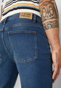 Τζιν σε σκούρο μπλε χρώμα με ανοιχτότερο ραβδισμό, διαθέτοντας μια πίσω τσέπη και μια καφέ δερμάτινη ετικέτα που αναγράφει "DENIM PROJECT."