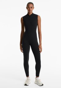 Mouwloze zwarte jumpsuit met een hoge halslijn en ritssluiting. Slanke pasvorm, gemaakt van een gladde, rekbare stof, gecombineerd met lichte sneakers.