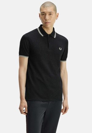 Joven con cabello corto que lleva una camiseta polo negra con ribete blanco en el cuello y las mangas, de pie frente a un fondo liso.