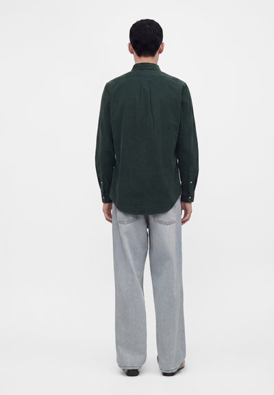 Homme aux cheveux courts et noirs, portant une chemise à manches longues vert foncé et un jean ample gris clair, debout de dos sur un fond blanc uni.