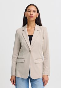 Beige sako s klopovými límci, jedním knoflíkem na zapínání a dvěma předními kapsami; hladká tkanina; přiléhavý design a dlouhé rukávy.