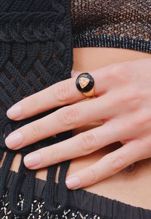 Bague dorée avec une pierre d'ambre triangulaire posée sur une base noire lisse, portée sur une main avec des ongles rose clair, vue en gros plan.
