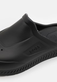 Chaussure noire en caoutchouc type slip-on avec un bout rond et une semelle texturée ; présente un logo en relief sur le côté et une surface lisse, conçue pour le confort.