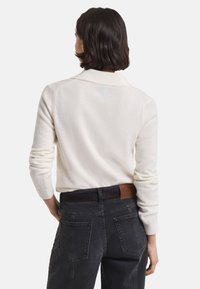 Vrouw met kort donker haar, gekleed in een witte langmouwen trui en zwarte high-waisted jeans, staat met haar rug naar de camera.