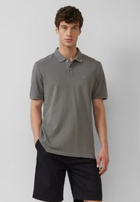 Grijze polo shirt van zacht materiaal, met een kraag, drie knopen, korte mouwen en een subtiel logo op de borst. Draagt met donkere shorts.