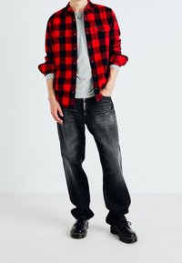 Rot-schwarz karierte Flanellhemd über einem grauen T-Shirt, kombiniert mit dunkelgrauen Jeans und schwarzen Schnürboots. Schlichtes Design mit funktionalen Taschen.