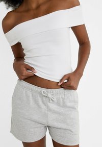 Witte off-shoulder top van rekbaar materiaal, gecombineerd met lichtgrijze shorts met trekkoord die een zachte textuur en een ontspannen pasvorm hebben.