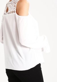 Blouse blanche avec design épaules dénudées et accents en dentelle au niveau du col. Texture douce et manches fluides, associée à un pantalon noir.