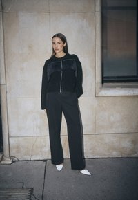 Schwarzer Pullover mit Fransen und Reißverschluss, kombiniert mit schwarzen weit geschnittenen Hosen und spitzen weißen Schuhen, vor einem neutralen Hintergrund.