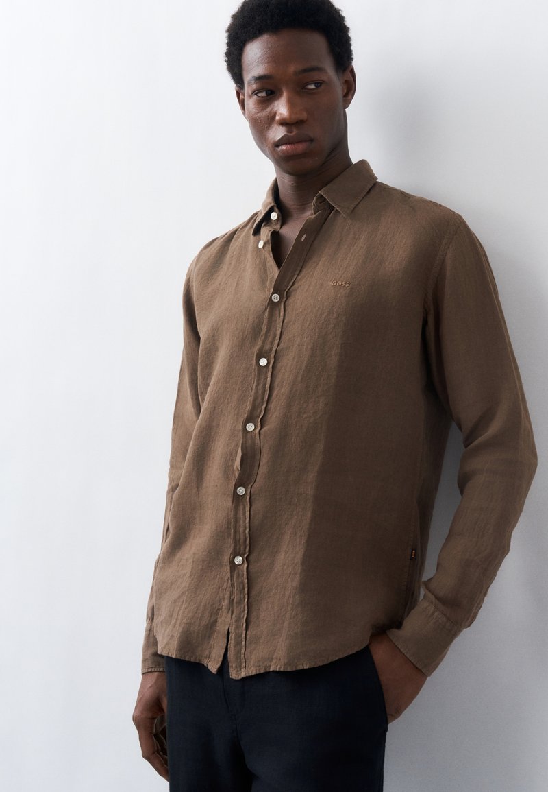 Άνδρας με καφέ πουκάμισο μακρυμάνικο με κουμπιά και σκούρο παντελόνι στέκεται με το ένα χέρι στην τσέπη, κοιτάζοντας ελαφρώς προς το πλάι, σε λευκό φόντο.