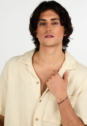 Camicia a maniche corte, color beige chiaro, con tessuto strutturato, abbottonatura frontale e taschino sul petto. Il modello indossa un bracciale al polso.