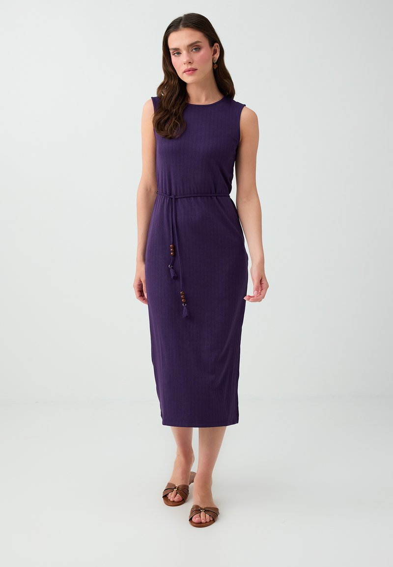 Femme aux longs cheveux foncés portant une robe midi violette sans manches avec une fine ceinture à la taille et des sandales marron à bout ouvert, debout sur un fond uni.