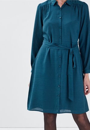 Woman wearing a teal button-up dress with long sleeves and a tied waist belt, standing against a plain light background.