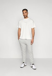 T-shirt branca de algodão com um pequeno logótipo preto, combinada com calças de treino cinza claro e sapatilhas brancas com detalhes em preto e dourado.