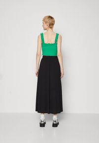 Zöld, ráncolt crop top fodros pántokkal, amelyet egy lenyűgöző fekete maxi szoknyával viselnek. A modell telitalpú ezüst cipőt és fehér zoknit hord.