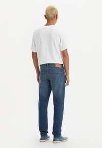 Camiseta blanca de algodón combinada con jeans de mezclilla azules, que presentan un acabado desgastado, diseño clásico de cinco bolsillos y ligero distressing. Calzado casual visible.
