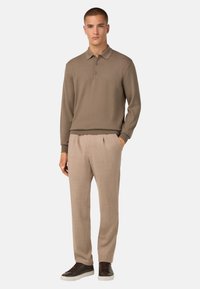 Hnědá polo košile s dlouhým rukávem a límečkem, se třemi knoflíky, v kombinaci se světle béžovými kalhotami na míru a hnědými teniskami. Čistý, texturovaný vzhled.