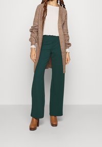 Lång beige kavaj i texturerad stickning, tillsammans med en vit ribbad topp och gröna vida byxor. Bruna klacktofflor fulländar looken.