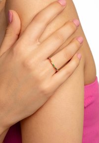 Anello in oro con gemme multicolori, caratterizzato da una fascia sottile e pietre distribuite in modo uniforme. La mano poggia su un braccio nudo con una pelle morbida.