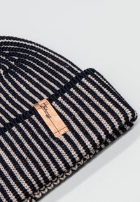 Cappellino a maglia con righe orizzontali blu navy e beige, caratterizzato da un tessuto testurizzato e da un'etichetta in pelle cucita lateralmente.