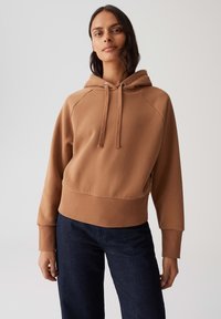 Vrouw in een bruine hoodie met trekkoorden en donkere spijkerbroeken, staand tegen een effen lichtgrijze achtergrond.