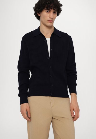 Cardigan bleumarin cu model canalat, cu guler, cu închidere butonată și mâneci lungi. Purtați peste o cămașă albă, asortat cu pantaloni bej deschis.