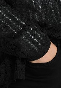 Černý krajkový svetr s texturovaným vzorem a kovovými akcenty, žebrovanými manžetami a volným střihem přes tmavý materiál.
