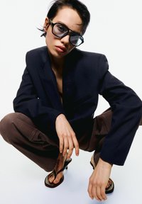 Black blazer with a notched lapel, brown wide-leg pants, oversized sunglasses, and brown flip-flops; model poses in a crouched position.