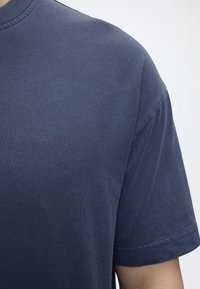 Κοντινό πλάνο ενός ατόμου που φοράει ένα μονόχρωμο σκούρο μπλε κοντομάνικο μπλουζάκι, δείχνοντας τον ώμο και το πάνω μέρος του μπράτσου σε λευκό φόντο.