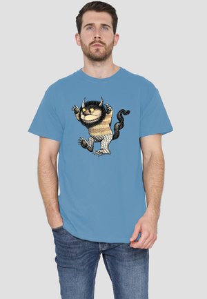 Mann trägt ein blaues T-Shirt mit einem Cartoon-Monster mit gestreiftem Körper, Hörnern, Krallen und einem gekrümmten Schwanz auf der Vorderseite.