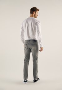 Chemise blanche à manches longues pour homme, jean gris avec poches arrière et baskets noires à semelles blanches. Fond neutre.