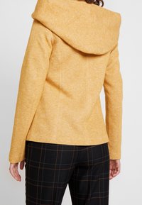 Sennepsgul, tekstureret stofjakke med en stor, foldet hætte og skræddersyet pasform, kombineret med sorte og brune ternede bukser.