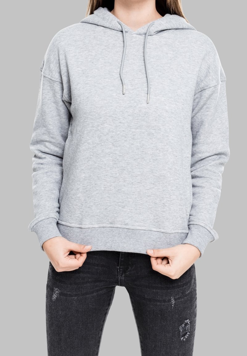 Mujer vestida con una sudadera gris claro y jeans negros desgastados, sosteniendo el dobladillo de la sudadera con ambas manos frente a un fondo gris.