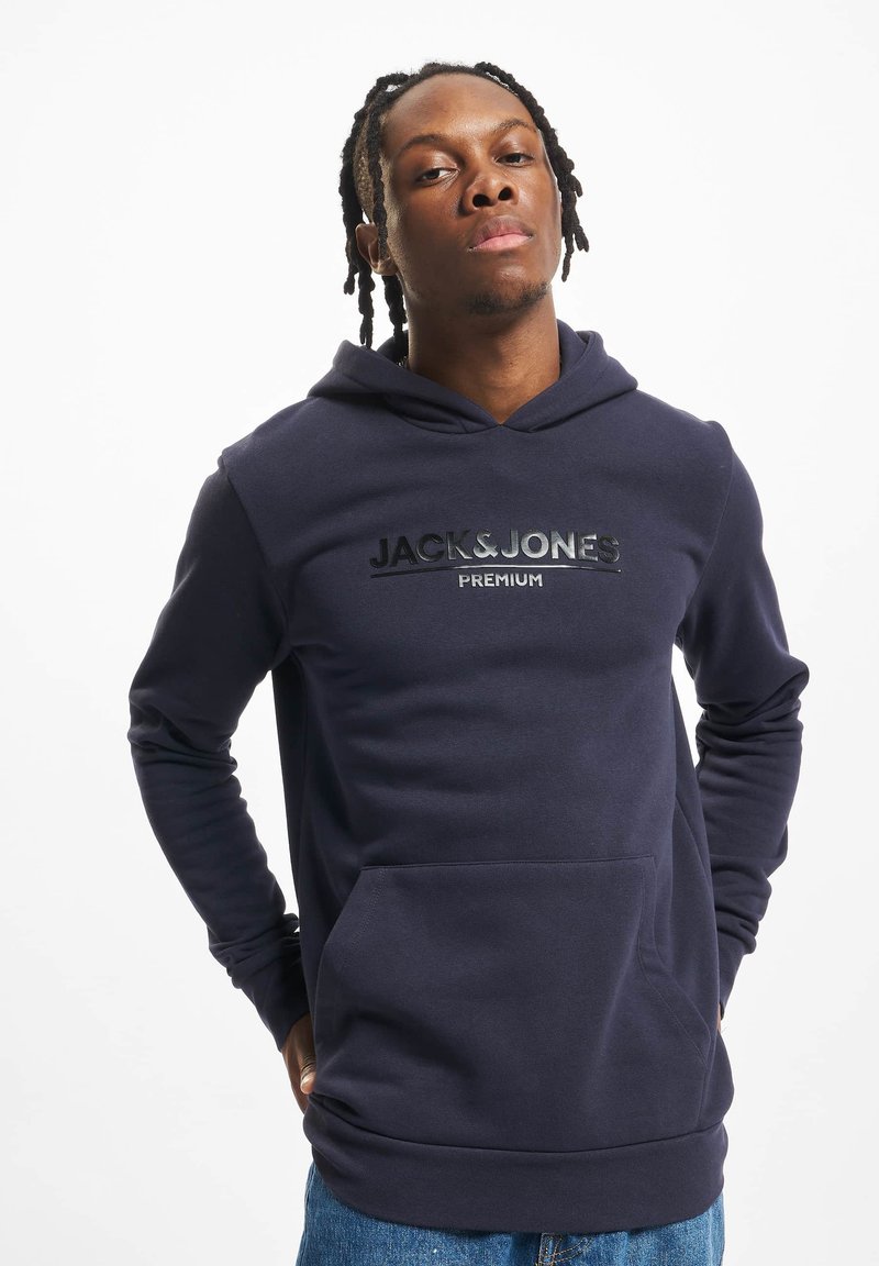 Ung mand med fletninger iført en marineblå Jack & Jones Premium hoodie og blå jeans, stående med hænderne bag ryggen.