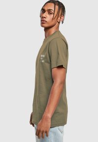 Oliven grøn t-shirt med korte ærmer lavet af bomuld, med teksten "ALDRIG TIL TID" i hvid på forsiden, rund hals og afslappet pasform.