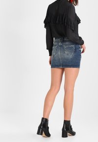 Femme portant un haut noir à manches longues et volants, une mini-jupe en denim bleu et des bottines noires à talons, se tenant de dos face à l'appareil photo.
