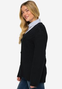 Cardigan nero a maglia con un motivo testurizzato a zigzag. Presenta un collo a V, bottoni frontali e maniche lunghe, abbinato a una camicia con colletto sotto.