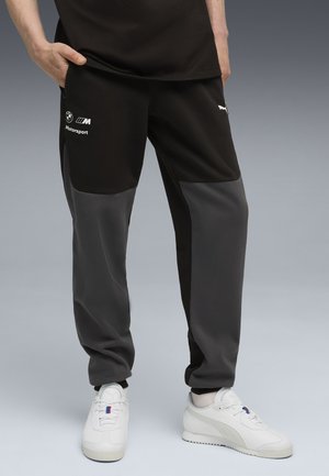 Pantaloni della tuta neri e grigi con un design affusolato, dotati di un logo e di testo sul lato. Realizzati in tessuto morbido con blocchi di colore a contrasto.