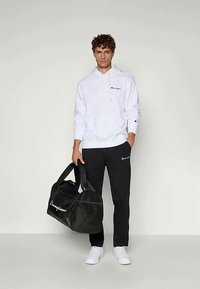 Sudadera blanca con un logo, pantalones negros con logo, y un bolso de deporte negro. El conjunto incluye zapatillas blancas. Fondo neutro.