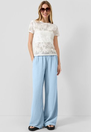 Frau mit großen runden Sonnenbrillen, weißem blumigem Spitzen-Kurzarm-Top, lockeren hellblauen Hosen und schwarzen Sandalen, stehend mit den Händen in den Taschen.