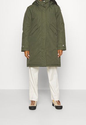 Person iført en olivengrøn hætteparka, hvide bukser og beige snørestøvler mod en ensfarvet hvid baggrund.