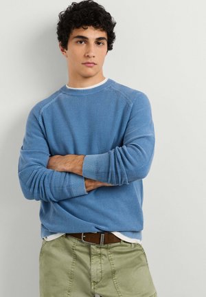 Junger Mann mit lockigem dunklem Haar, der einen blauen strukturierten Pullover und olivgrüne Hosen trägt, steht mit verschränkten Armen vor einem hellen Hintergrund.