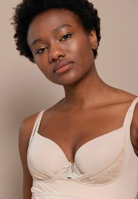 Reggiseno beige in pizzo con spalline regolabili, caratterizzato da un design della coppa liscia, accenti in satin e delicate rifiniture in pizzo al centro.