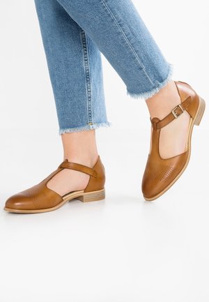 Brune læder T-rem sko med perforerede detaljer båret med lyseblå jeans med frynsede kanter på hvid baggrund.