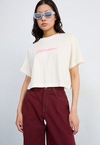Avklippt krämfärgad t-shirt med rosa broderad text, i kombination med vida bourgognebyxor. Modellen bär genomskinliga ljusblå solglasögon.