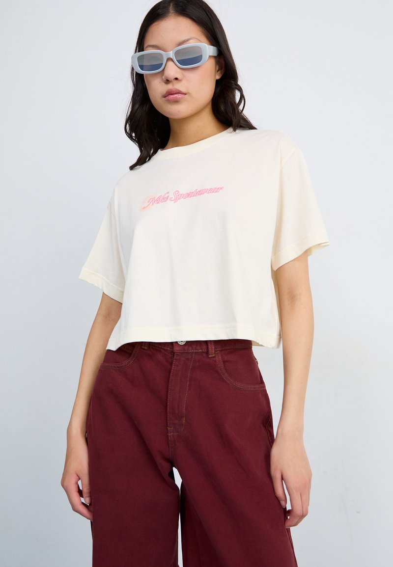 Camiseta de crema recortada con texto bordado en rosa, combinada con pantalones anchos burdeos. El modelo lleva gafas de sol translucidas de color azul claro.