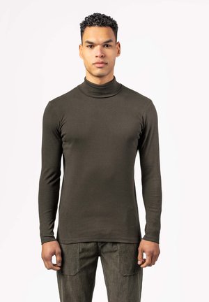 Young man with short curly hair wearing a fitted dark olive turtleneck and matching pants, standing against a plain light background.