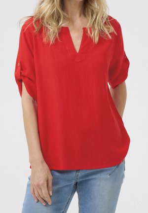 Mujer con una blusa roja suelta con las mangas remangadas y vaqueros azul claro frente a un fondo liso.