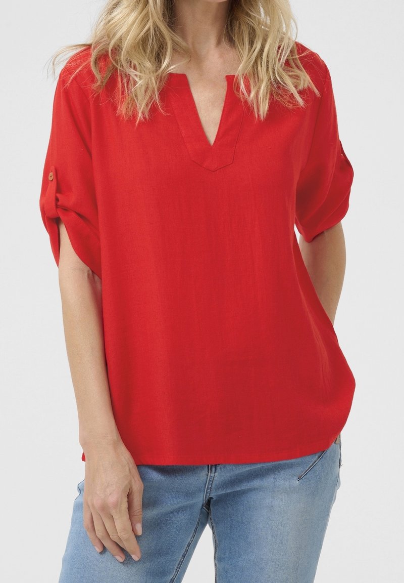 Femme portant un chemisier rouge ample aux manches retroussées et un jean bleu clair sur un fond uni.