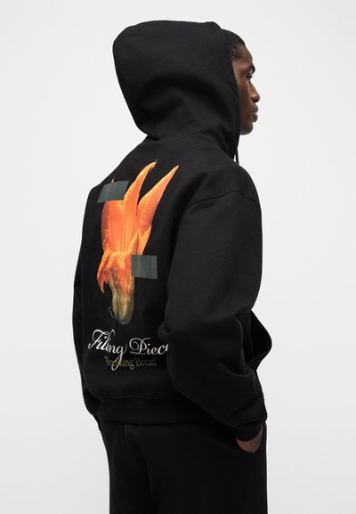 Άνδρας που φοράει μαύρο φούτερ με κουκούλα, η οποία καλύπτει το κεφάλι του, με πορτοκαλί αφηρημένο σχέδιο και το κείμενο «Filing Pieces Breaking Bread» στην πλάτη.